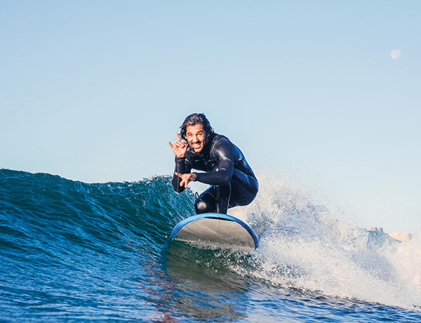 Surfer crouching on a wave making a shaka hand sign, moon visible in daytime sky