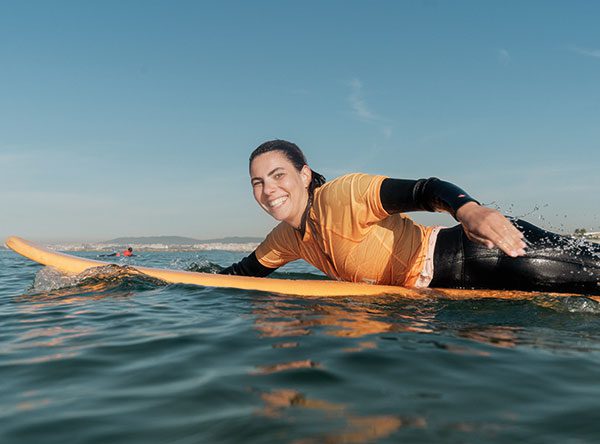 Woman paddling on an orange surfboard smiling at the camera with coastline in the distance