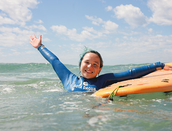 Young surfer in a Gota Dagua wetsuit waving from the water at our kids surf camp