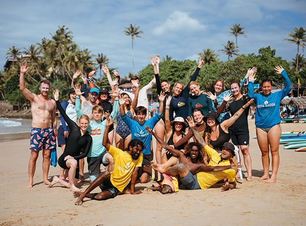 Group of surf campers cheering with raised arms on a palm-lined beach in Sri Lanka