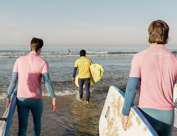 Three surfers in rash guards carrying boards while walking into the ocean