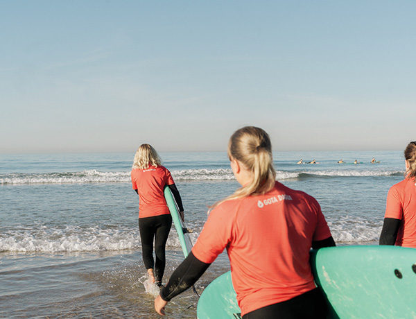 Students in red rash guards carrying boards into the ocean on student surf trips