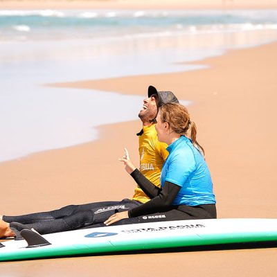 Surf student and instructor sitting on the beach next to a surfboard, laughing together