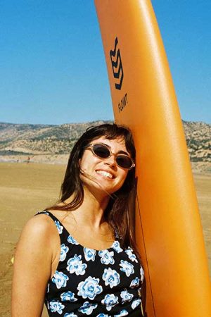 Woman in sunglasses leaning against an orange surfboard on a sandy beach