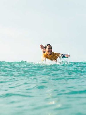 Surfer in a yellow rash guard laughing while paddling through turquoise water