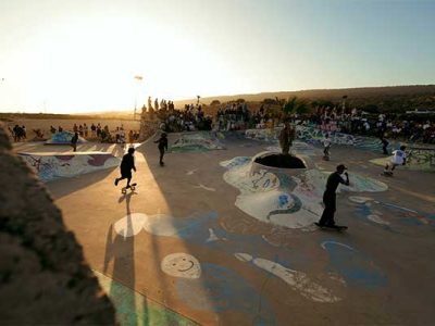 Skateboarders riding a sunlit outdoor skate bowl as a crowd looks on