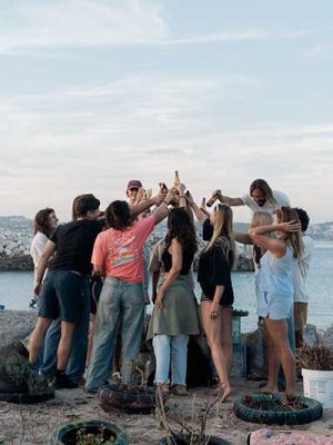 Friends toasting drinks together at sunset on a rocky shoreline by the sea