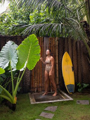Woman smiling at an outdoor bamboo shower beside a yellow surfboard, surrounded by tropical plants