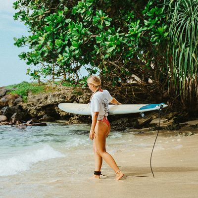 Woman carrying a surfboard along a rocky beach shaded by tropical trees