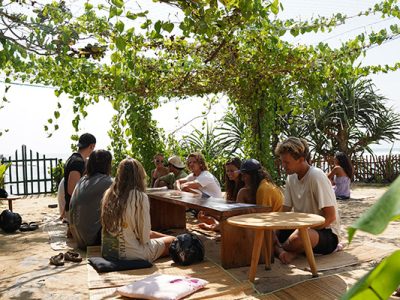 Group of surfers relaxing around a low table under a leafy pergola near the beach