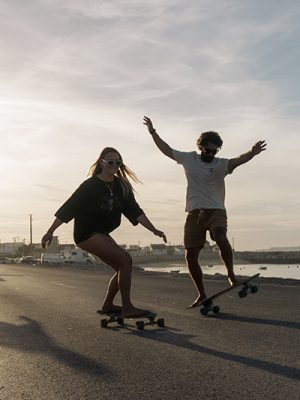 Two people longboarding along a coastal road at sunset, one with arms raised