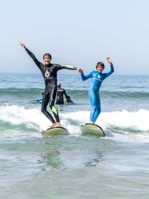 Two kids in Gota Dagua wetsuits riding a wave together, holding hands